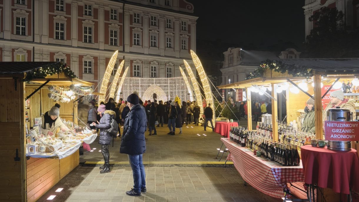 Zdjęcie przedstawia jarmark na pl. Kolegiackim. Na pierwszym planie stoiska handlowe, w tle grupę ludzi oglądających świąteczne iluminacje.