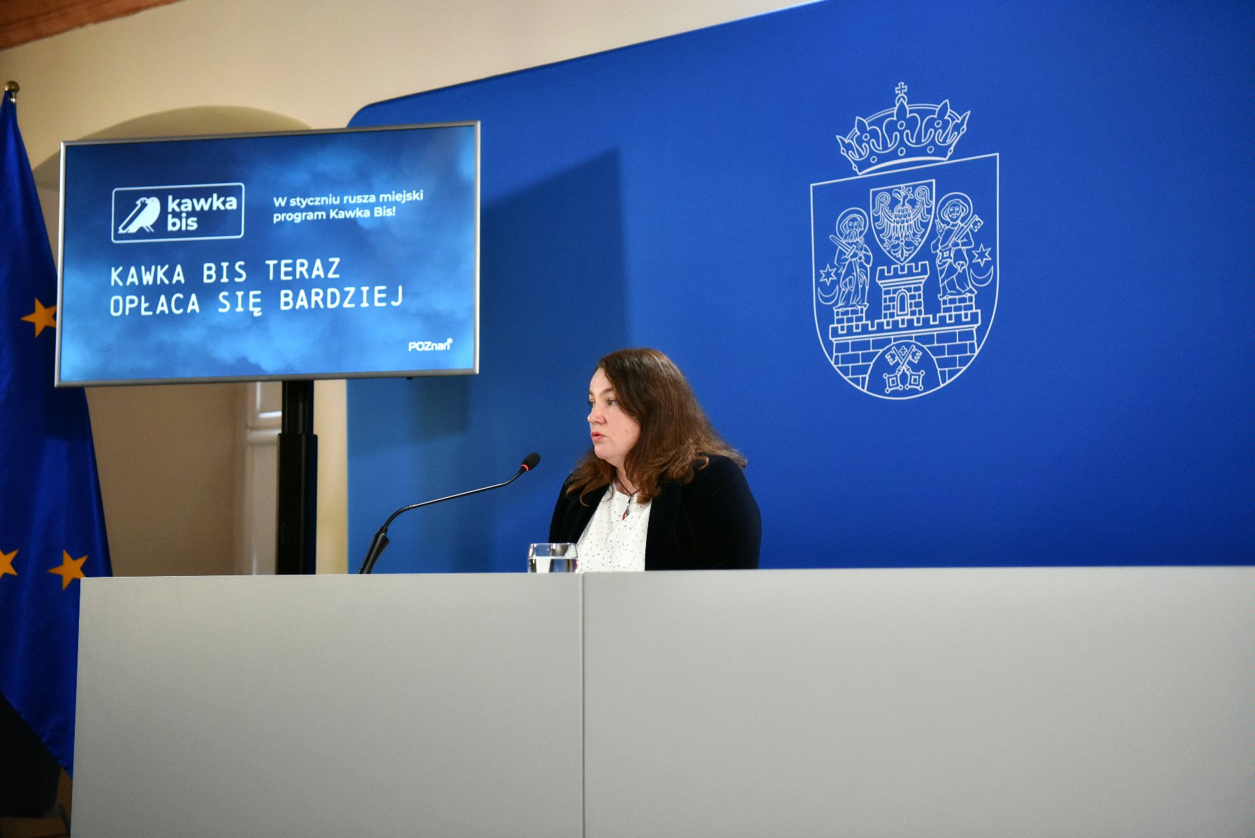Na zdjęciu kobieta siedząca za stołem i mówiąca do mikrofonu, za nią niebieskie tło i telewizor - grafika artykułu