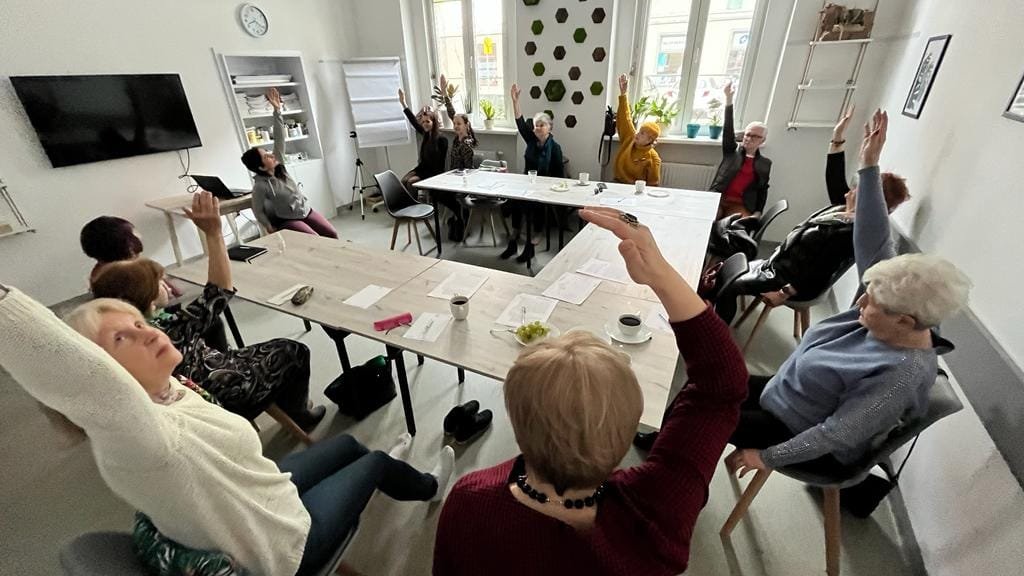 Na zdjęciu grupa ludzi przy stole, każdy z nich ma podniesioną w górę rękę