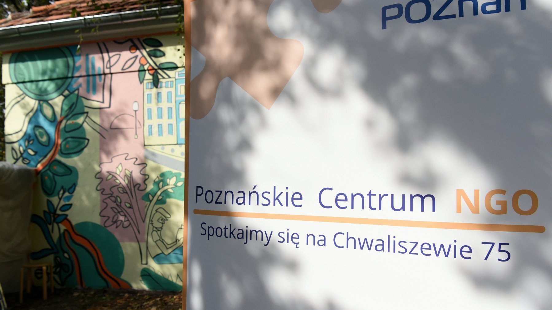 Plakat Poznańskiego Centrum NGO, w tle mural na ścianie kamienicy