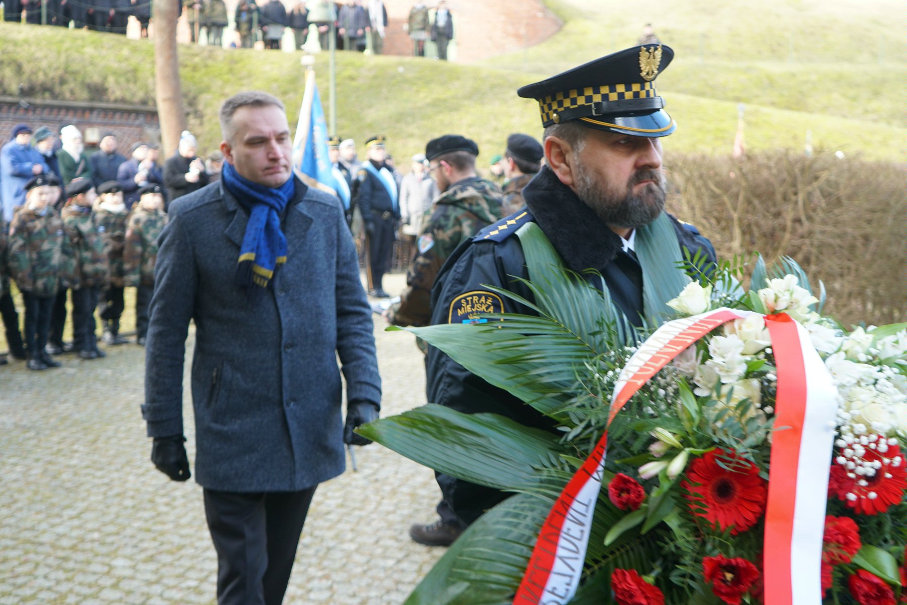 Na zdjęciu zastępca prezydenta Poznania składajacy kwiaty, przed nim strażnik miejski z wiązanką - grafika artykułu