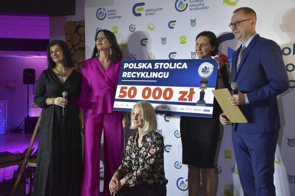 Na zdjęciu piątka ludzi w eleganckich strojach trzymająca bon na 50 000 złotych - grafika artykułu