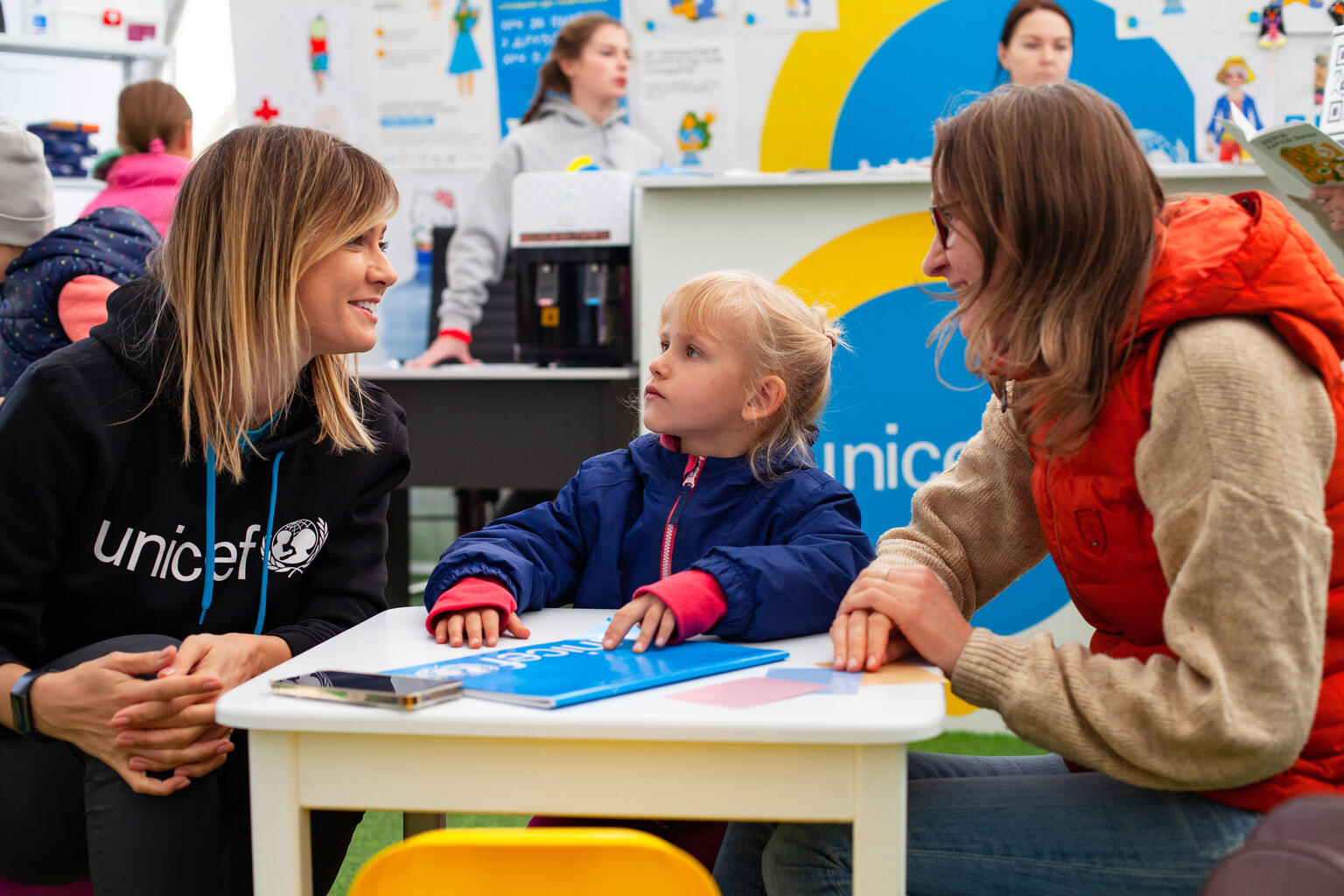 Na zdjęciu dwie kobiety i dziewczynka przy małym stole, jedna z kobiet ma bluzę z logotypem UNICEF - grafika artykułu