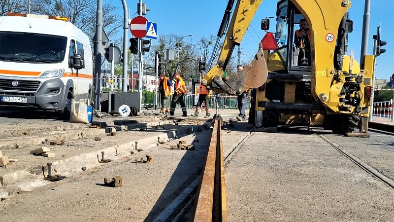 Zdjęcie przedstawia pracowników służb torowych układającyh torowisko. W tle koparka.