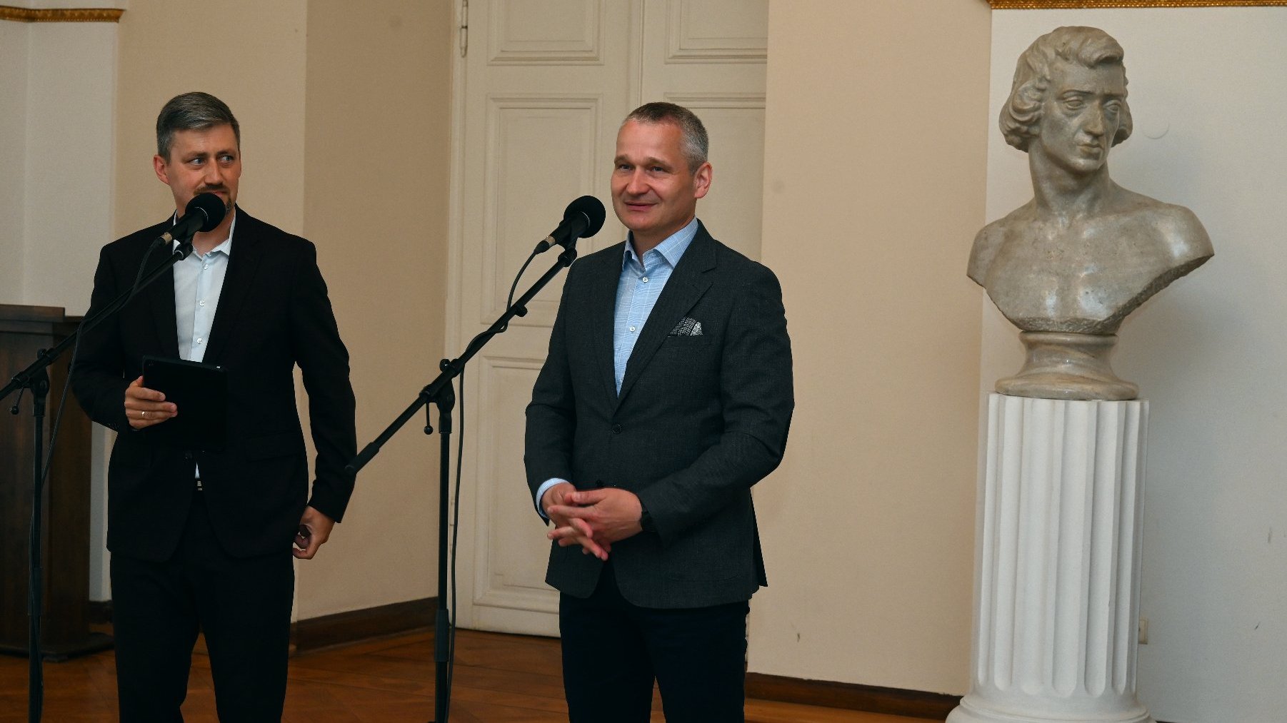 Na zdjęciu dwóch mężczyzn przed mikrofonami, za nimi stoi rzeźba popiersia Chopina