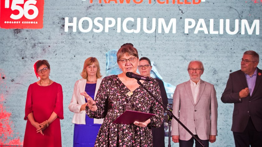 na zdjęciu kobieta mówiąca coś do mikrofonu, za nią stoi kilka osób, w tle napis: Hospicjum Palium