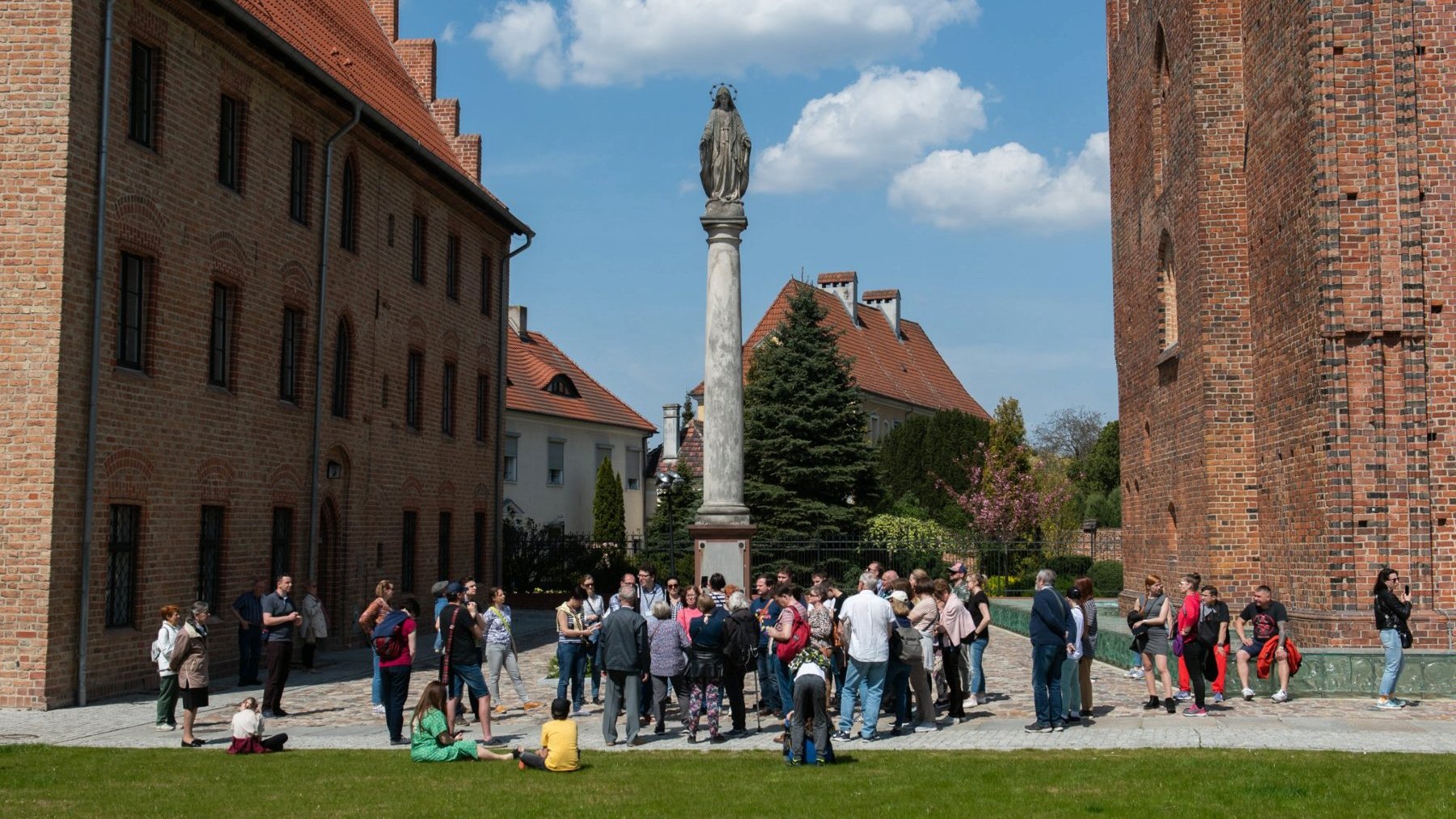 Zdjęcie przedstawia grupę ludzi podczas spaceru po Ostrowie Tumskim. W tle ceglane mury budynków oraz monument zwieńczony figurą.