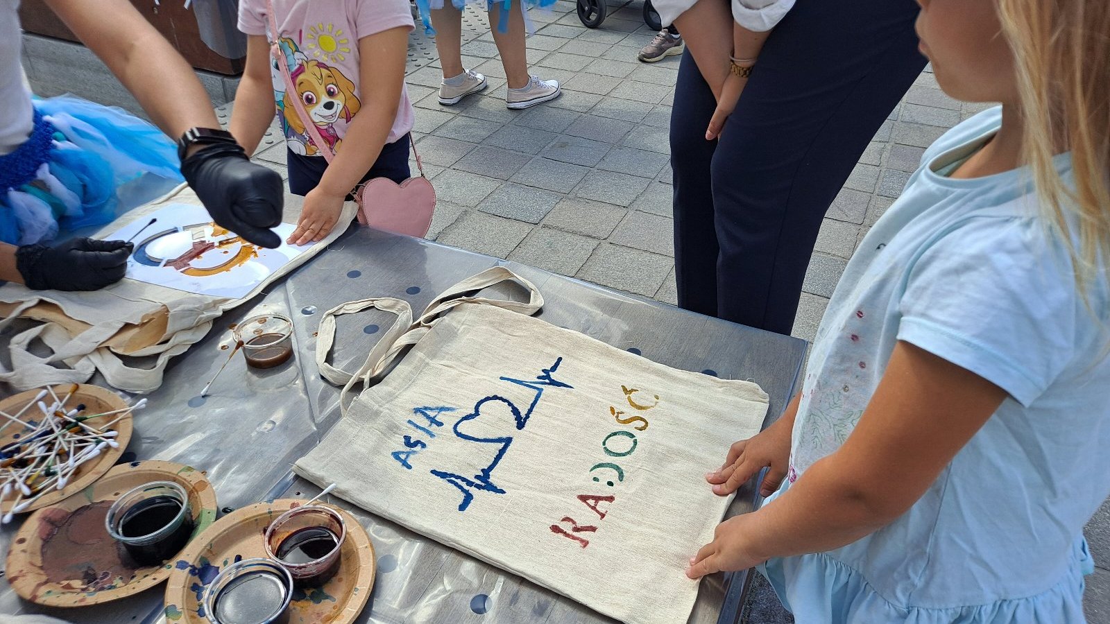 Na zdjeciu dzieci tworzące rękodzieło, dziewczynka trzyma dłonie na torbie z napisem: Asia, Radość