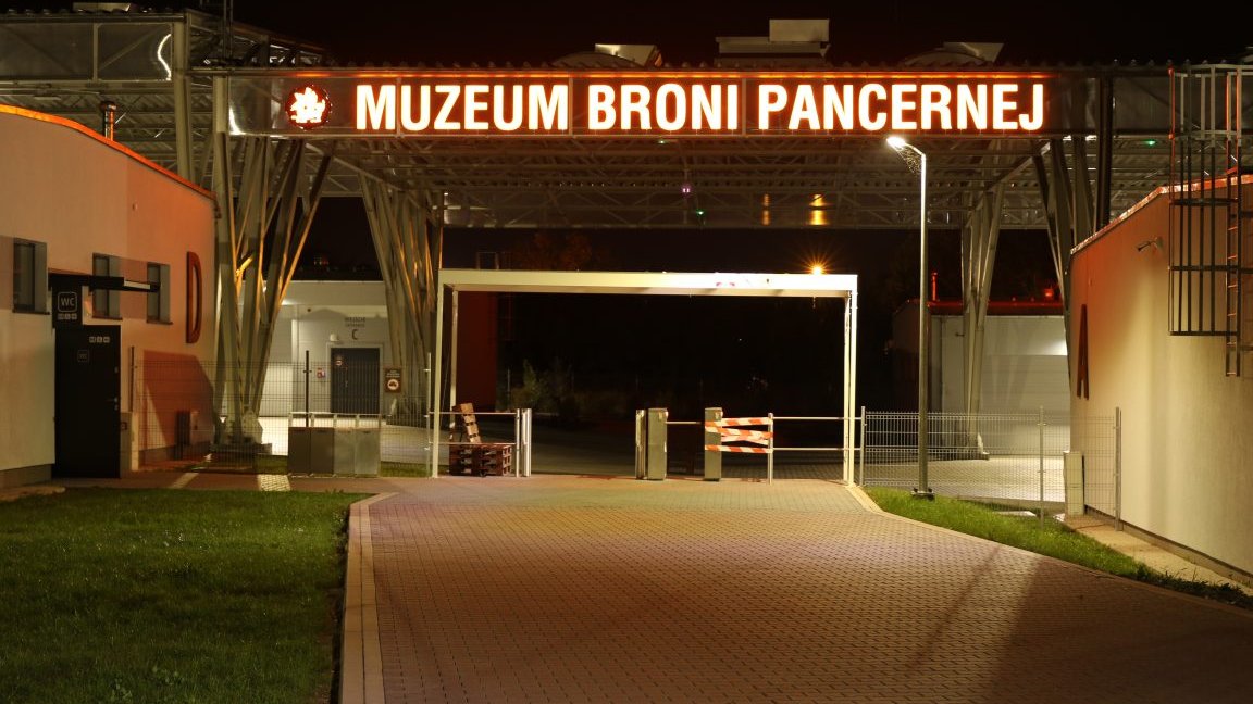 Zdjęcie sprzed wejścia do muzeum broni pancernej - widać napis oraz bramkę i budynki