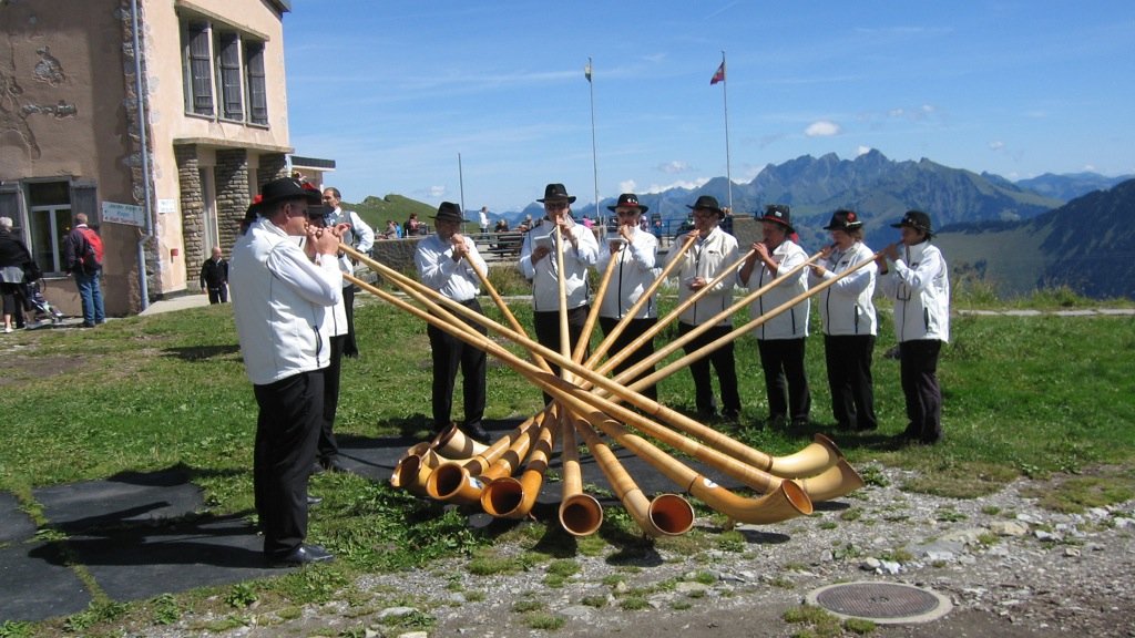 Zdjęcie muzyków grających na dużych rogach, w tle Alpy