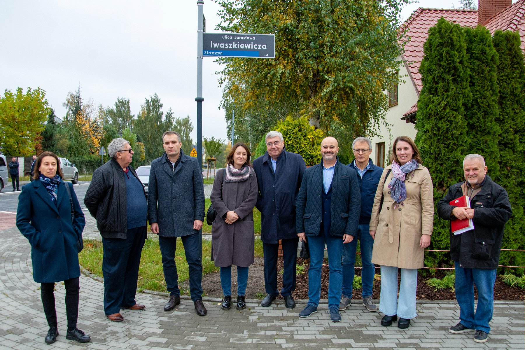 Zdjęcie przedstawia grupę osób pozujących do zdjęcia. W tle widać zieleń, drogę oraz tablicę z nazwą: ulica Jarosława Iwaszkiewicza. - grafika artykułu