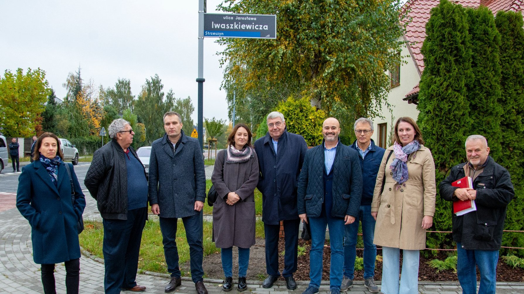 Zdjęcie przedstawia grupę osób pozujących do zdjęcia. W tle widać zieleń, drogę oraz tablicę z nazwą: ulica Jarosława Iwaszkiewicza.