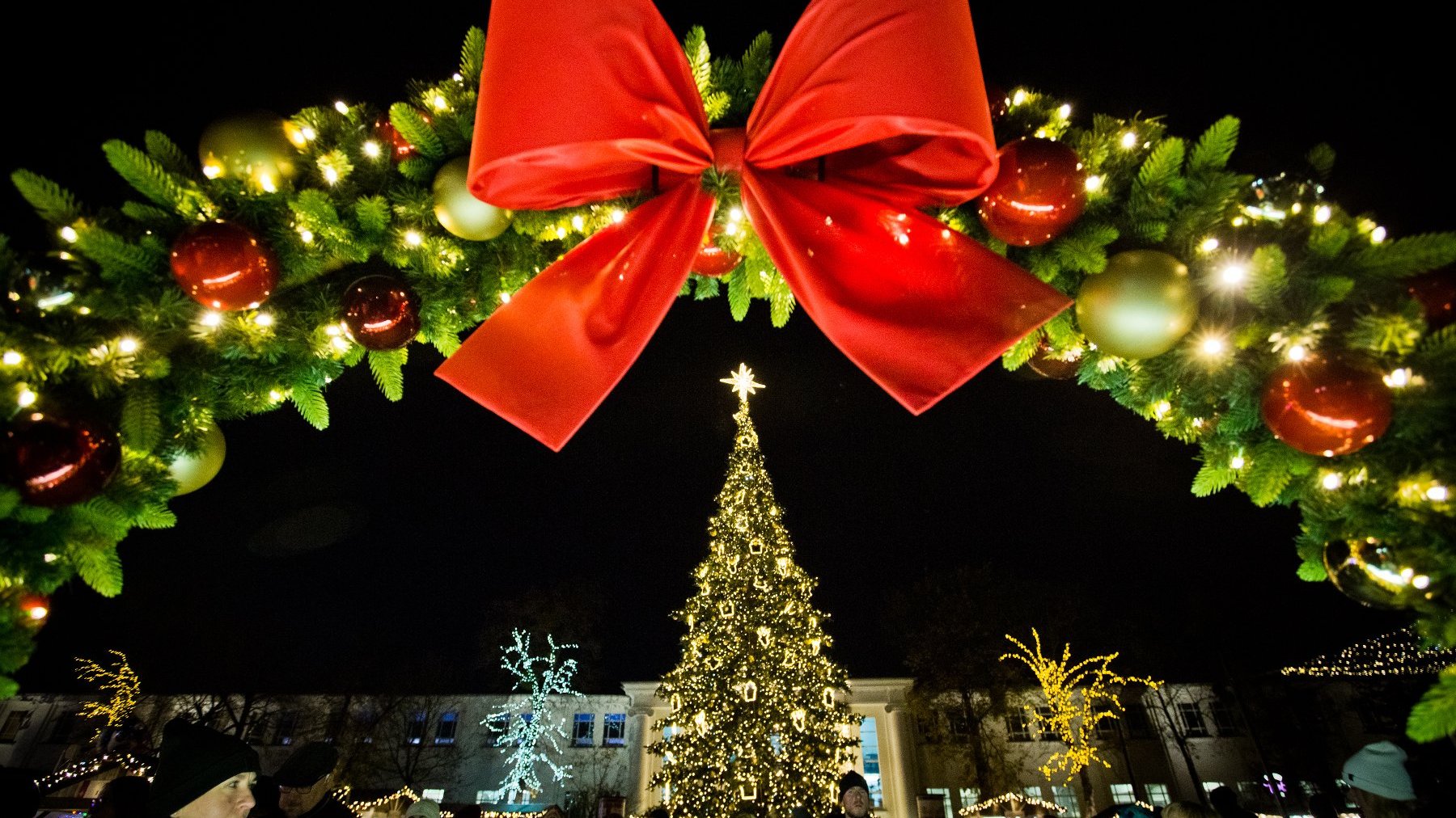 W centrum kadru widać udekorowaną lampkami choinkę, powyżej zaś wielki świąteczny wieniec z kokardą.