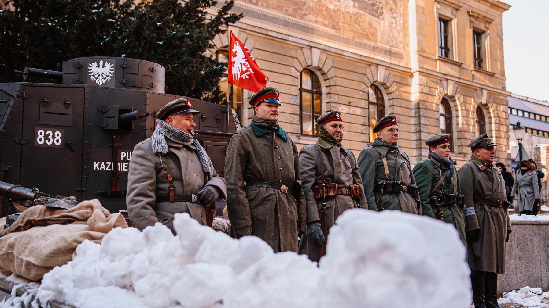 Zdjęcie przedstawia grupę rekonstruktorów w mundurach Powstania Wielkopolskiego. Za nimi stoi pojazd opancerzony.