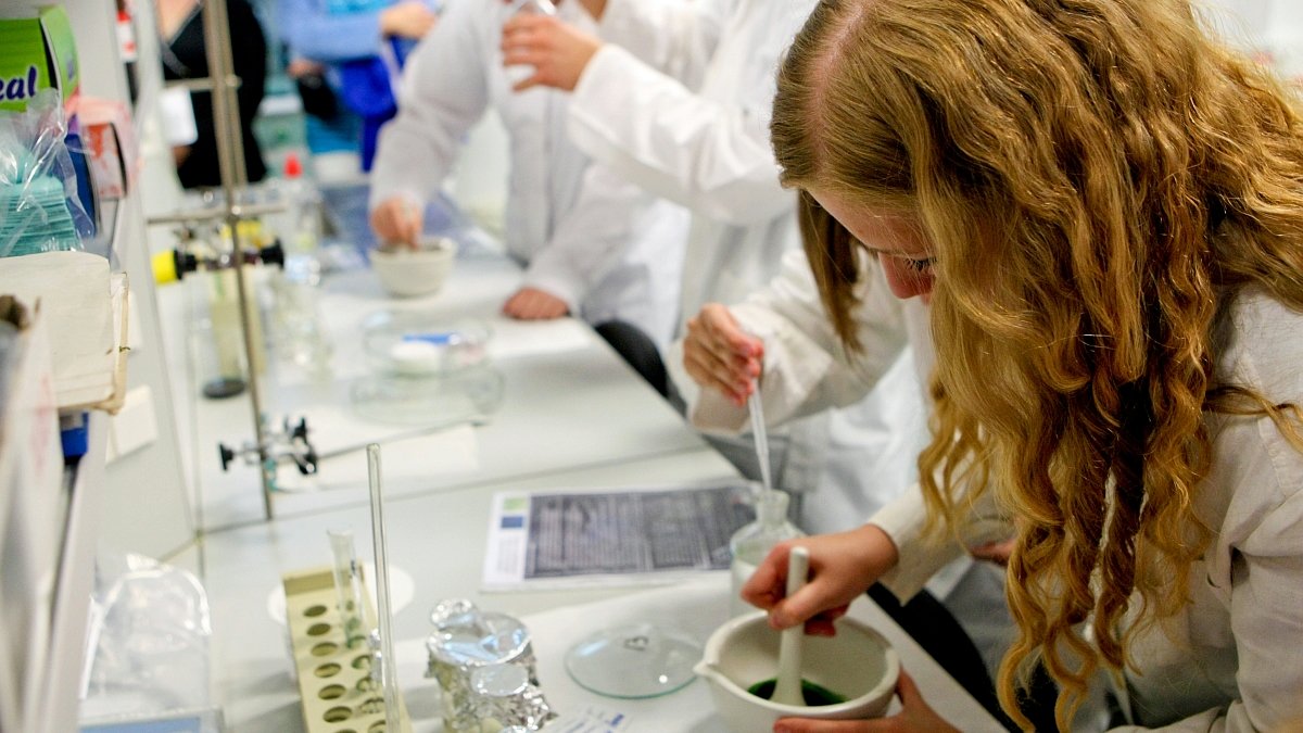 Zdjęcie przedstawia osoby w białych fartuchach w laboratorium. Na pierwszym planie znajduje się dziewczyna mieszająca jakąś substancję w pojemniku. - grafika artykułu