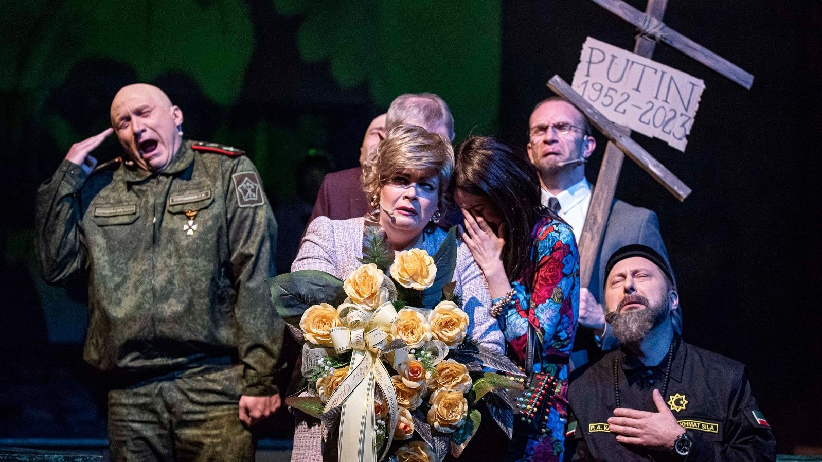 Zdjęcie przedstawia płaczących i zmartwionych ludzi. Jeden z mężczyzn trzyma drewniany krzyż z tabliczką z napisem "Putin 1952-2023".