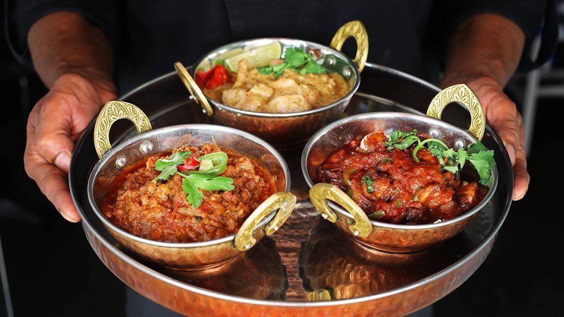 Na zdjęciu dłonie trzymające tacę, na niej trzy miski z hinduskimi daniami