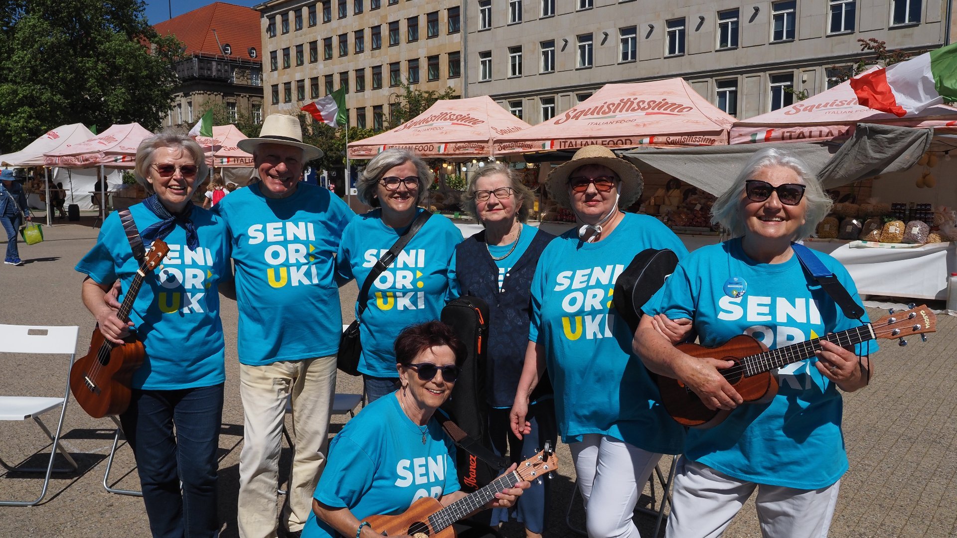 Na zdjęciu uśmiechnięciu seniorzy i seniorki w niebieskich koszulkach, każdy trzyma w ręku ukulele