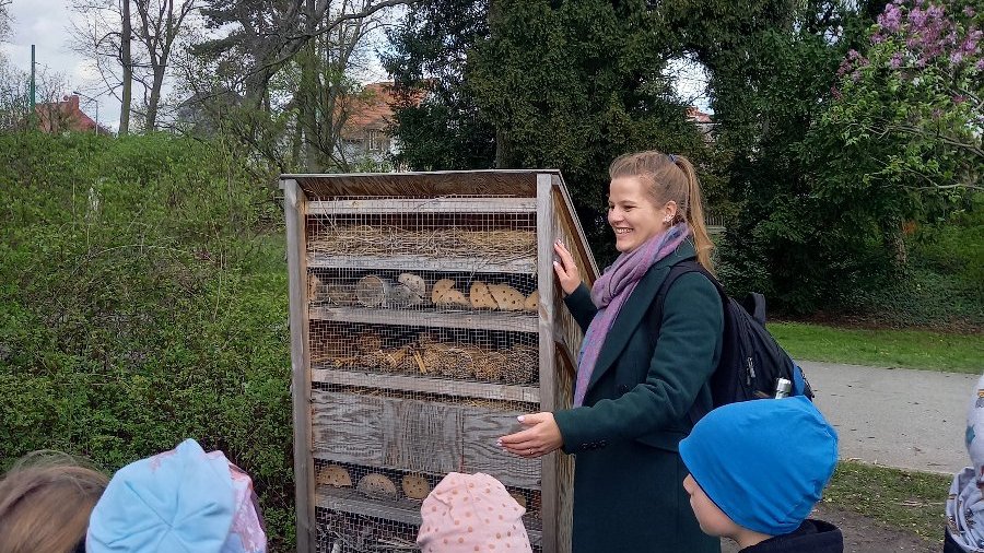 Zdjęcie przedstawia kobietę prezentującą dzieciom domek dla owadów.