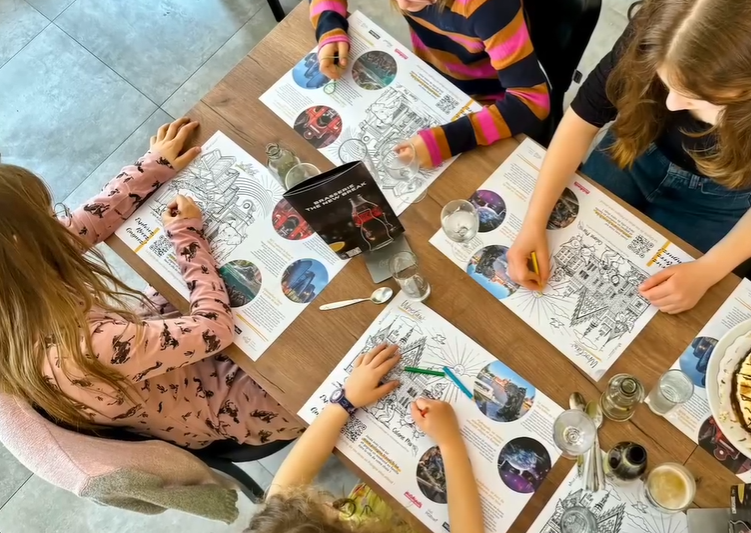 Na zdjęciu stolik widziany z góry, na nim kolorowanki, przy nim dzieci - grafika artykułu