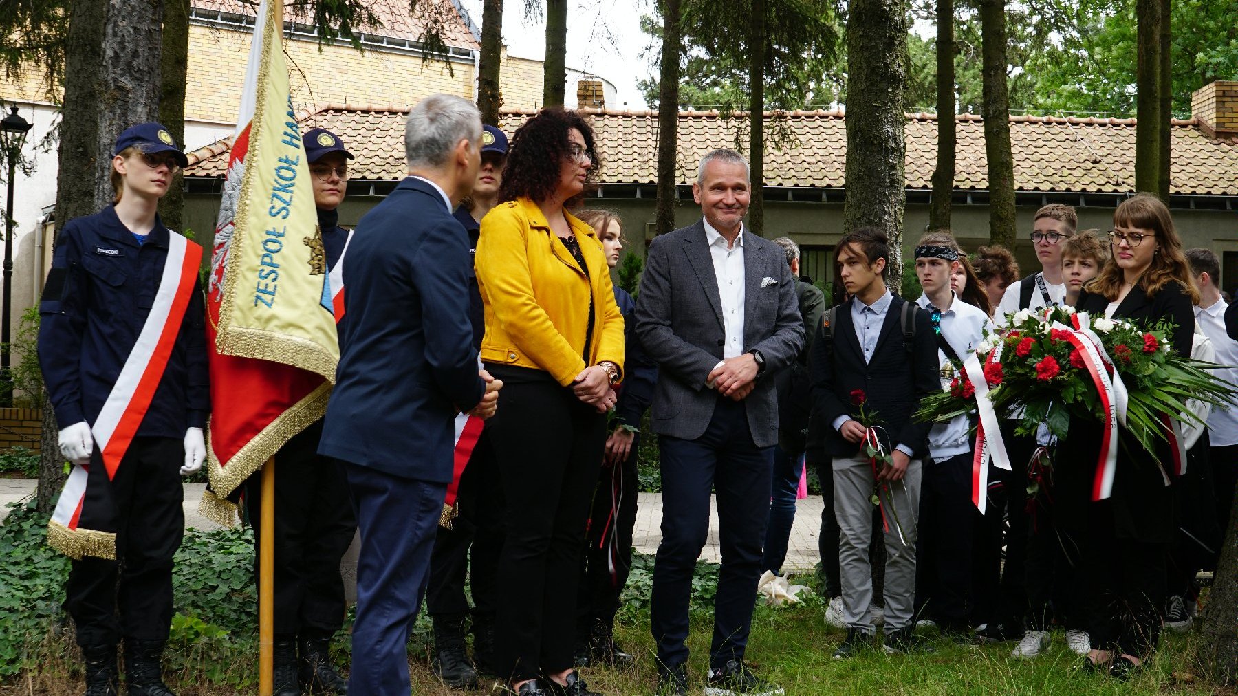 Zdjęcie przedstawia uczniów i dorosłych z kwiatami i sztandarami.