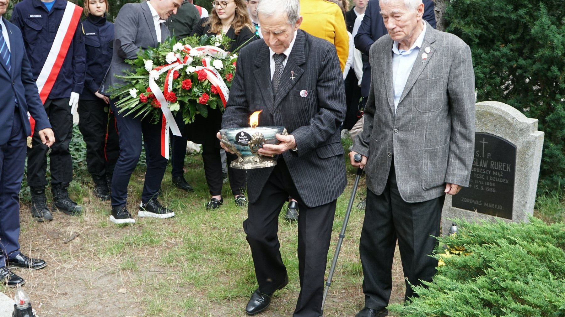 Zdjęcie przedstawia dwóch starszych mężczyzn idących z zapalonym zniczem.