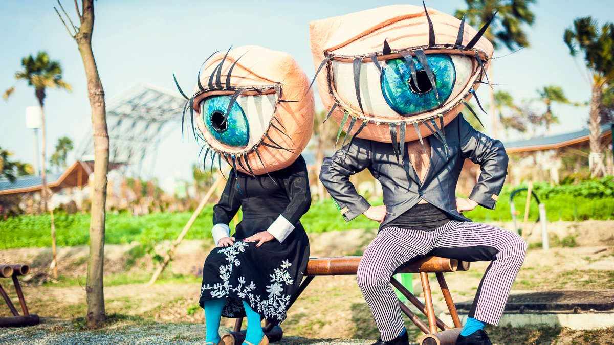 Zdjęcie przedstawia dwójkę ludzi siedżących na ławce w maskach w kształcie oka.