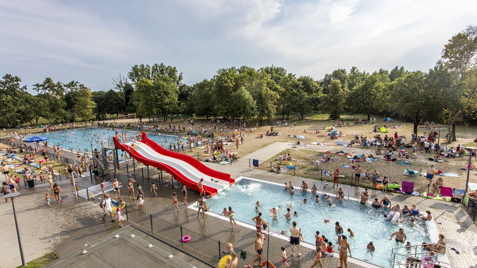 Zdjęcie przedstawia tłum ludzi na pływalni letniej, składającej się z dwóch basenów i zjeżdżalni.