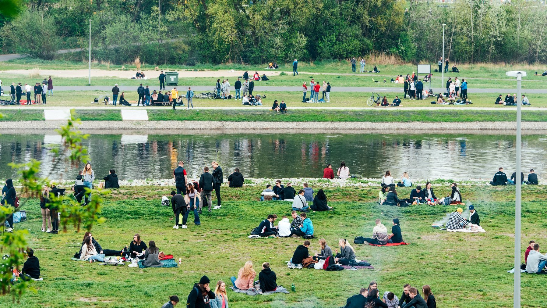 Zdjęcie przedstawia ludzi nad rzeką. Niektórzy siedzą na kocach, inni stoją i rozmawiają.