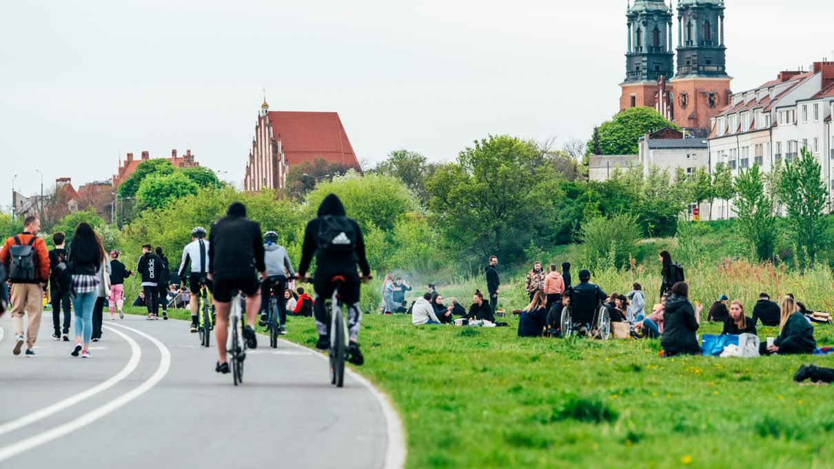 Zdjęcie przedstawia ludzi na teranach nadwarciańskich. Niektórzy siedzą na kocach, inni spacerują lub jeżdzą na rowerach. W tle widać Katedrę Poznańską.