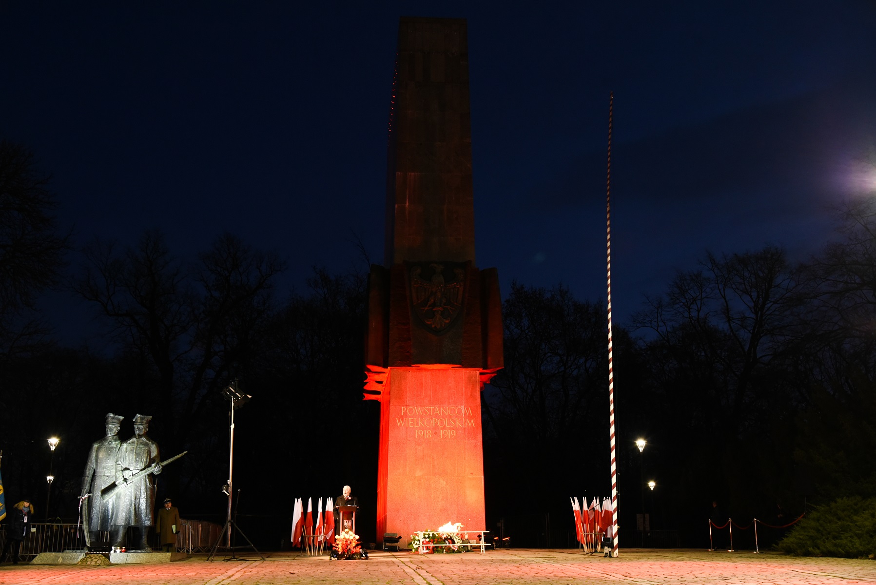 Obraz przedstawia pomnik oświetlony w nocy. Pomnik jest poświęcony Powstańcom Wielkopolskim z lat 1918-1919, o czym świadczy napis 'POWSTAŃCOM WIELKOPOLSKIM 1918-1919' na frontowej części. Pomnik jest podświetlony na czerwono, a po jego lewej stronie znajduje się statua dwóch żołnierzy. Przed pomnikiem znajduje się kilka flag Polski oraz osoba stojąca za podium, prawdopodobnie wygłaszająca przemówienie lub prezentację. - grafika artykułu