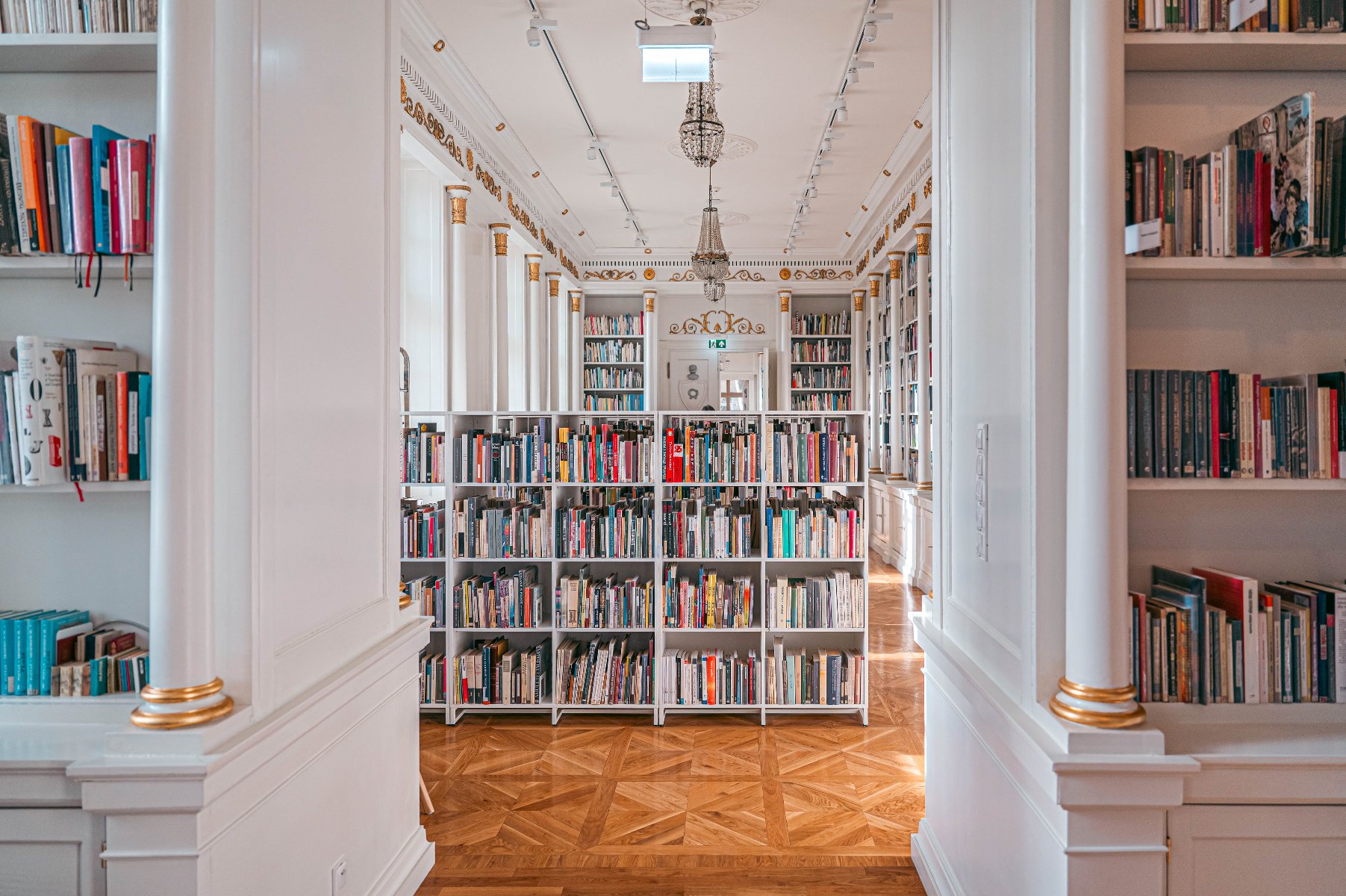 Zdjęcie przedstawia wnętrze dobrze zorganizowanej biblioteki. Biblioteka ma wysokie białe regały wypełnione różnorodnymi książkami. Regały są ustawione w rzędach, tworząc przejścia, które ułatwiają dostęp do książek. Pomieszczenie ma wysoki sufit z ozdobnymi dekoracjami i żyrandolami, co dodaje eleganckiego charakteru wnętrzu. Podłoga jest wykonana z polerowanego drewna z geometrycznym wzorem. Biblioteka jest jasno oświetlona, a naturalne światło wpada przez duże okna po prawej stronie pokoju. - grafika artykułu