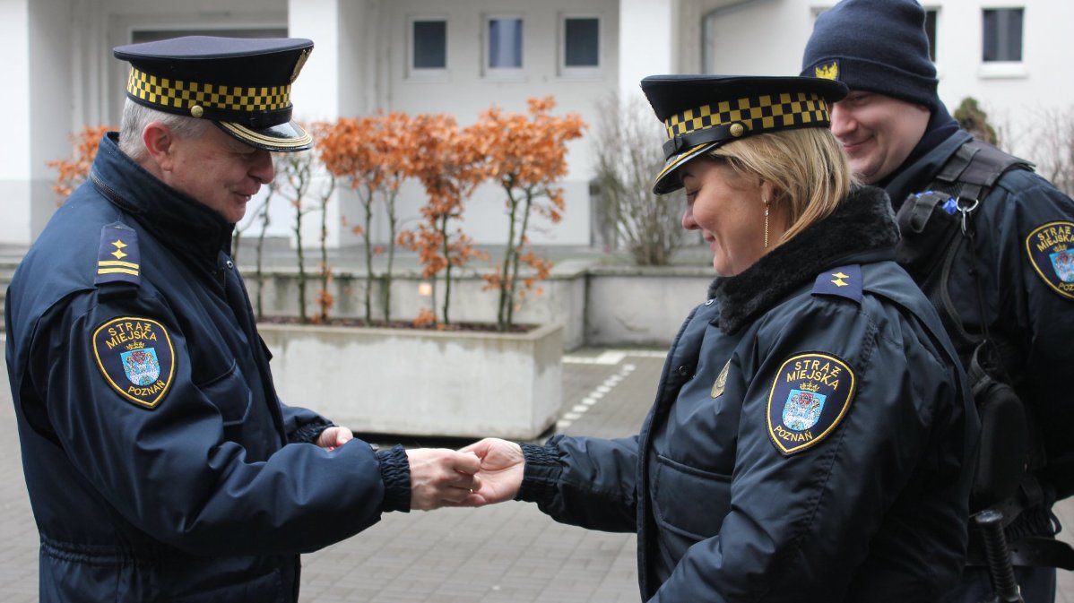 Na obrazie widzimy trzech funkcjonariuszy Straży Miejskiej Poznań stojących na dziedzińcu. Ubrani są w mundury z odznakami i naszywkami, które wskazują na ich przynależność do straży miejskiej. Funkcjonariusze stoją obok siebie, a jeden z nich wręcza dokument drugiemu. W tle widoczne są rośliny oraz budynek. Scena ma charakter oficjalny.