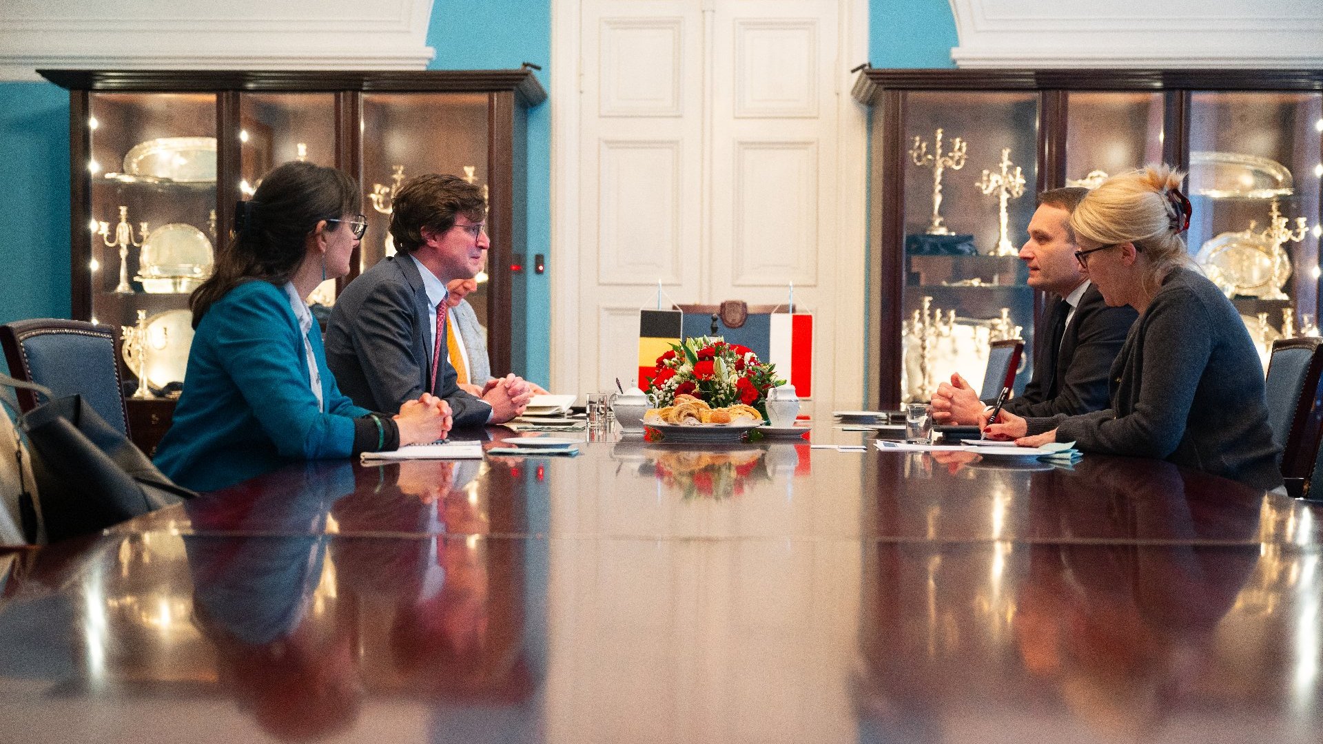 Na obrazku widzimy formalne spotkanie, prawdopodobnie w sali konferencyjnej. Przy dużym, drewnianym stole siedzą cztery osoby, po dwie z każdej strony. Stół ma wypolerowaną powierzchnię, a przed uczestnikami znajdują się notatniki, papiery i długopisy. Na środku stołu znajduje się dekoracyjna aranżacja z kwiatami i flagami, prawdopodobnie wskazująca kraje lub organizacje reprezentowane na spotkaniu.