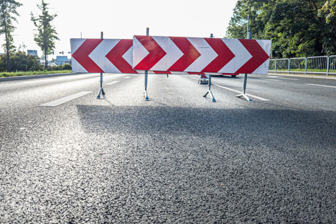 Na zdjęciu widać barierę na drodze wielopoziomowej. Bariera składa się z trzech metalowych podpór wspierających prostokątne tablice z czerwonymi i białymi wzorami szpiczaków wskazującymi w lewo. Droga znajduje się w obszarze miejskim, z widocznymi drzewami i budynkami w tle. Bariera prawdopodobnie służy do kierowania ruchem poza zamkniętą lub ograniczoną sekcję drogi. - grafika artykułu