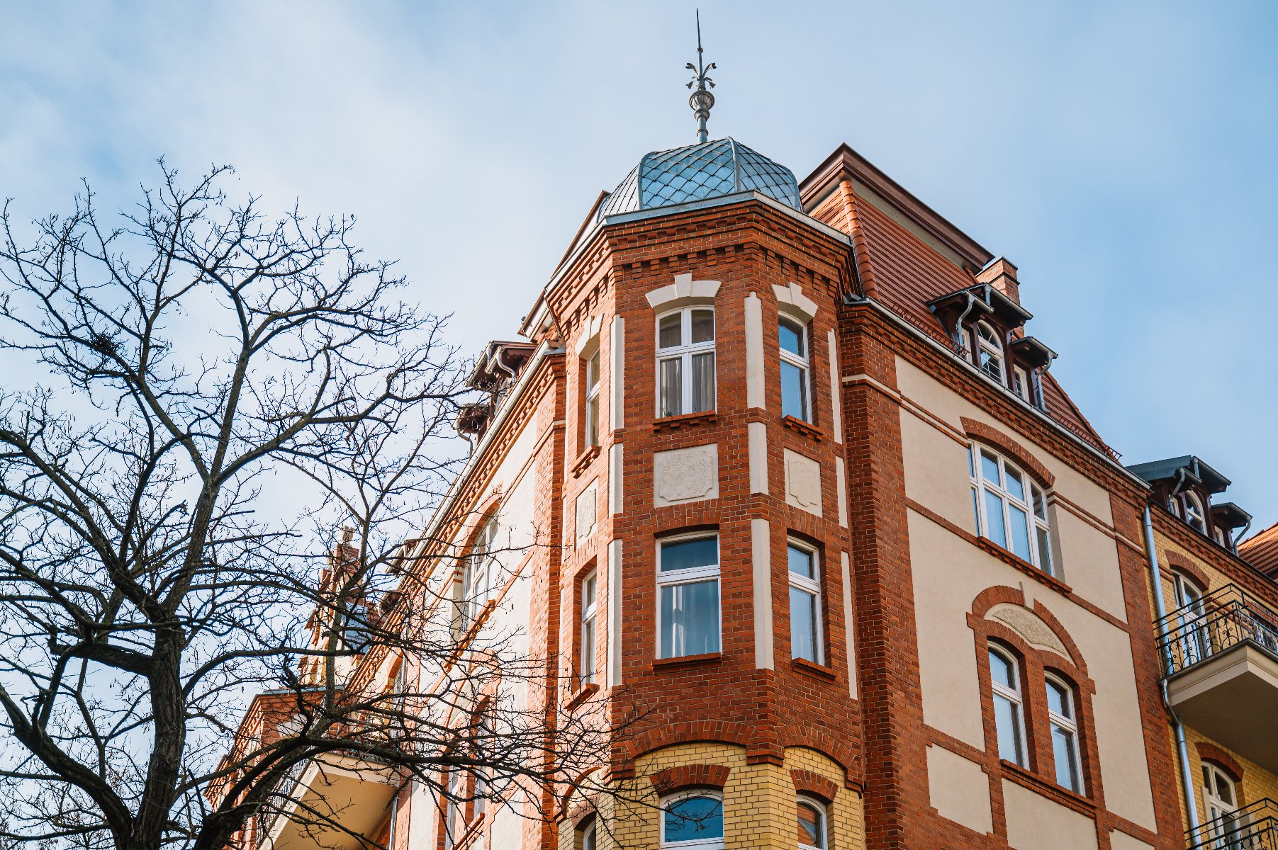 Budynkek mieszkalny o wielokondygnacyjnym stylu architektonicznym. Na zdjęciu widać połączenie czerwonej cegły z jasnym tynkiem oraz ozdobne detale wokół okien i linii dachowej. Dach ma stromy spadek z lukarnami i małą kopułą u góry. Widać kilka balkonów z kutymi żelaznymi balustradami. Na pierwszym planie znajduje się drzewo bez liści. - grafika artykułu