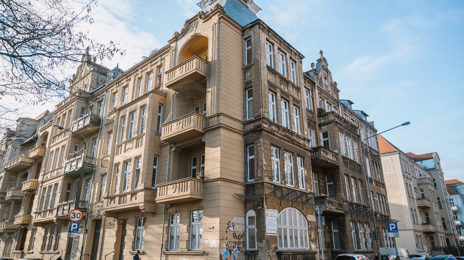 Widok uliczny budynku mieszkalnego o ozdobnym stylu architektonicznym. Budynek ma wiele kondygnacji z dużymi oknami oraz licznymi balkonami. Fasada udekorowana jest skomplikowanymi detalami, a dach ma okna połaciowe i lukarny. Na ulicy znajdują się zaparkowane samochody, a w pobliżu budynku widocznych jest kilka osób.