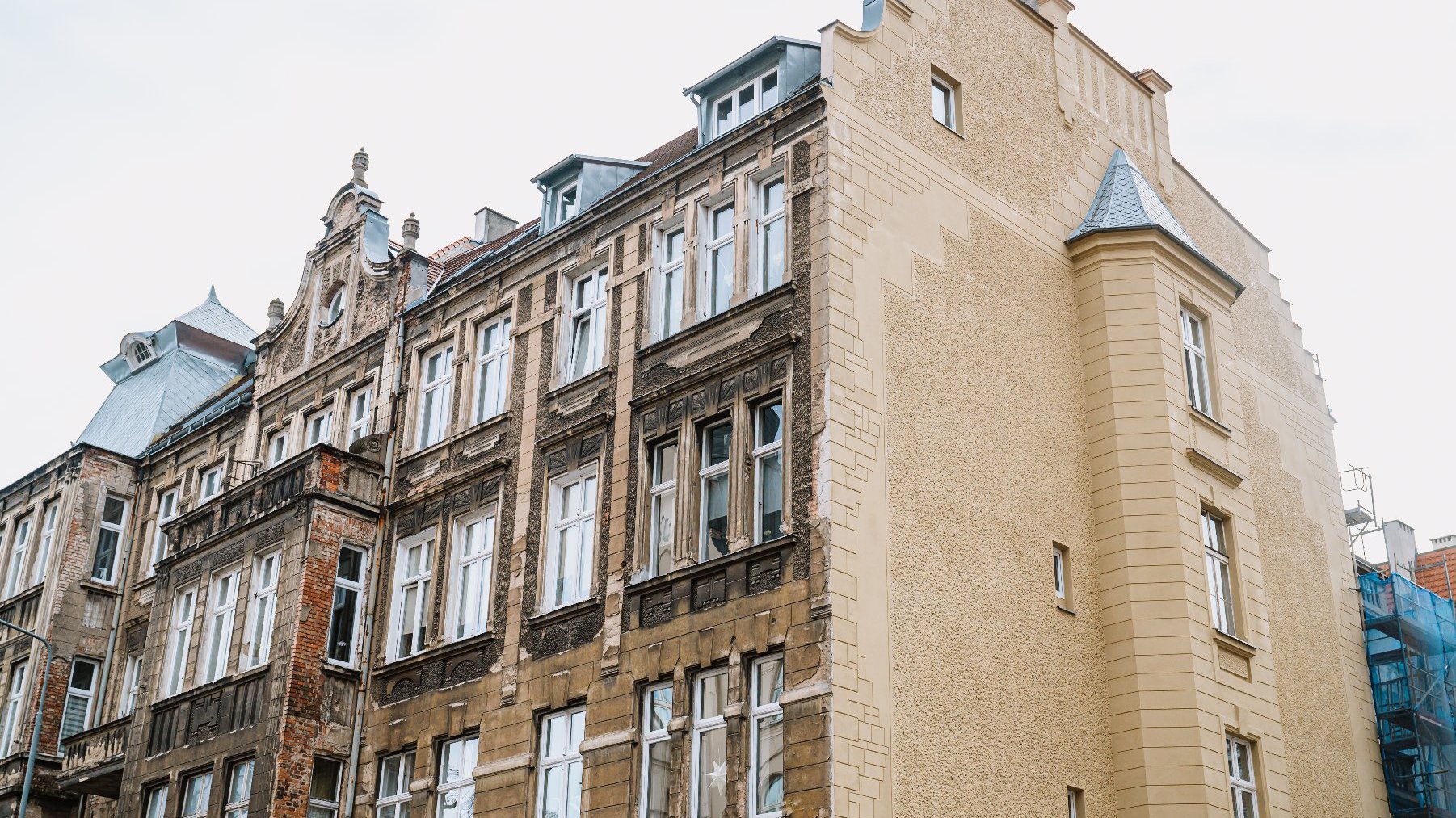 Obrazek ukazuje wielopiętrowy budynek o ozdobnym stylu architektonicznym. Budynek łączy elementy stare i nowe, z lewej strony wygląda na bardziej zniszczony i starzejący się, podczas gdy prawa strona wydaje się być niedawno odnowiona lub dobrze utrzymana. Strukturę budynku zdobią liczne okna, niektóre z nich są otwarte, a fasadę uzupełniają dekoracyjne elementy takie jak detale sztukatorskie oraz dach mansardowy. Prawa strona budynku ma charakterystyczny schodkowy szczyt i małą wieżyczkę z dachem stożkowym.