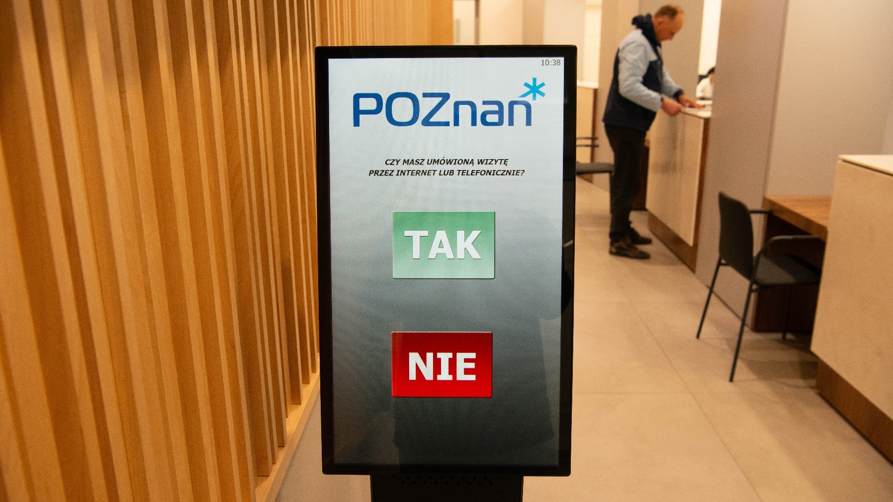 Ekran dotykowy w publicznym biurze obsługi klienta. Na ekranie widnieje tekst "POZnan" na górze, a pod nim pytanie: "CZY MASZ UMÓWIONĄ WIZYTĘ PRZEZ INTERNET LUB TELEFONICZNIE?" Poniżej znajdują się dwa duże przyciski: zielony oznaczony "TAK" i czerwony oznaczony "NIE". W tle widoczna jest osoba stojąca przy ladzie, prawdopodobnie rozmawiająca z przedstawicielem obsługi.