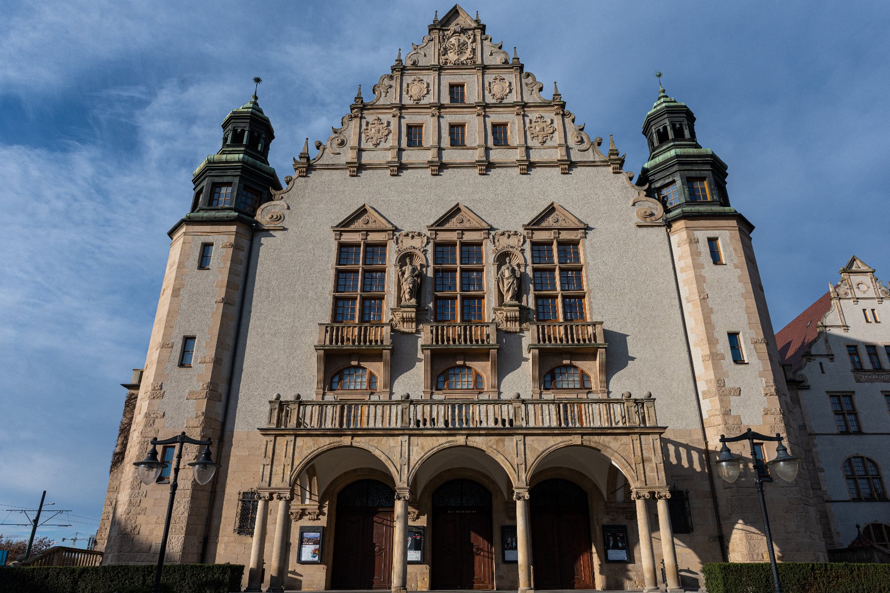 Duży ozdobny budynek o symetrycznej fasadzie. Budynek ma dwie wieże po bokach z zielonymi kopułowymi dachami. Centralna część budynku ma stromy dach z ozdobnymi elementami i posągami. Na dole znajdują się trzy łukowate wejścia, każde z drewnianymi drzwiami, a powyżej balkon. B - grafika artykułu