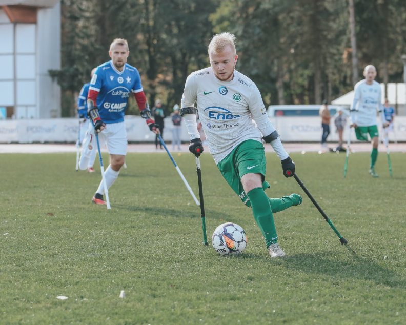 Grupa sportowców gra w amp futbol na boisku trawiastym. Zawodnicy poruszają się, używając kul. Na pierwszym planie gracz w białej i zielonej koszulce z numerem 20 kontroluje piłkę lewą stopą. Ma na sobie zielone spodenki i zielone getry. W tle widoczny jest inny zawodnik w niebiesko-białym stroju oraz kilku graczy w biało-zielonych strojach. W tle drzewa i budynek. - grafika artykułu