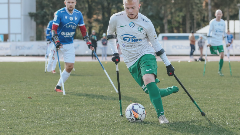 Grupa sportowców gra w amp futbol na boisku trawiastym. Zawodnicy poruszają się, używając kul. Na pierwszym planie gracz w białej i zielonej koszulce z numerem 20 kontroluje piłkę lewą stopą. Ma na sobie zielone spodenki i zielone getry. W tle widoczny jest inny zawodnik w niebiesko-białym stroju oraz kilku graczy w biało-zielonych strojach. W tle drzewa i budynek.