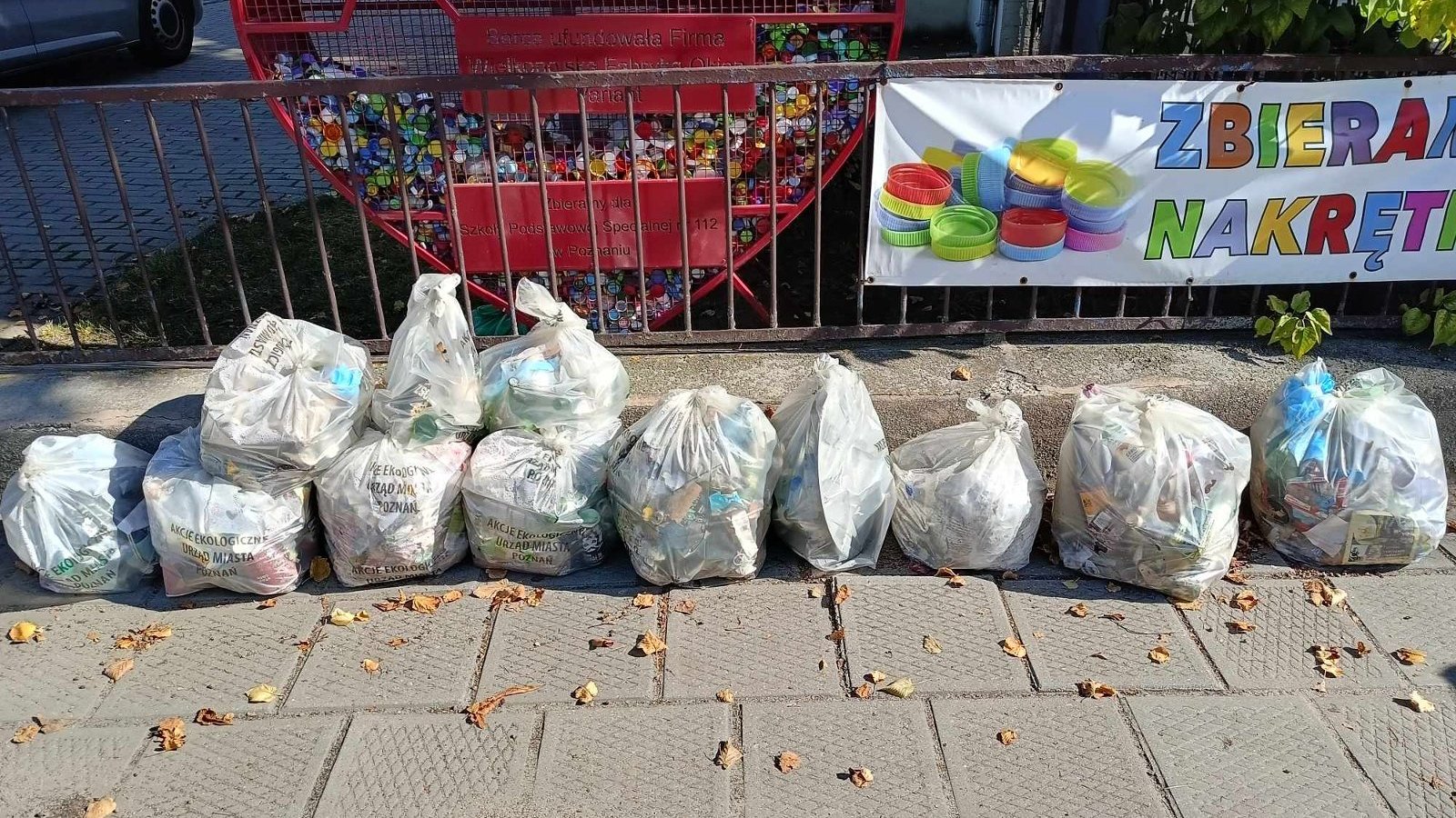 Zdjęcie przedstawia worki ze śmieciami zebrane w jednym miejscu. Śmieci leżą na chodniku. Z tyłu widać ogrodzenie i pojemnik na plastikowe nakrętki w kształcie serca.