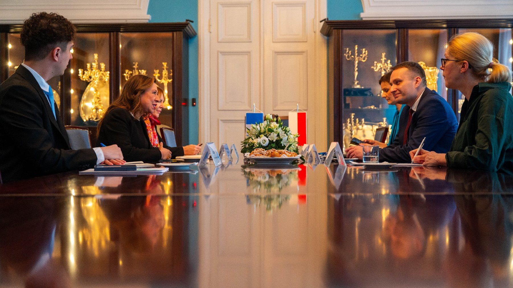 Formalne spotkanie odbywające się w elegancko udekorowanym pomieszczeniu. Przy dużym drewnianym stole siedzą cztery osoby, po dwie z każdej strony, twarzą do siebie. Na stole widoczne są tabliczki z nazwiskami i dokumenty, co sugeruje, że jest to spotkanie dyplomatyczne lub biznesowe. Na środku stołu znajduje się kompozycja kwiatowa oraz dwie małe flagi