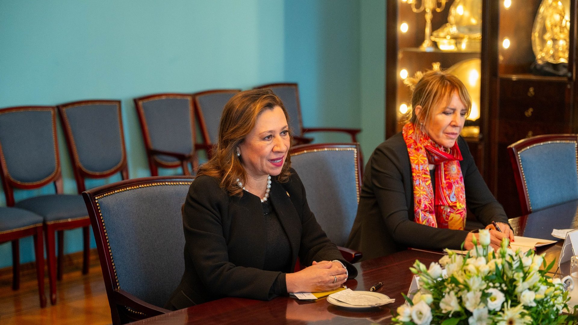 Formalne spotkanie w elegancko udekorowanym pomieszczeniu. Przy stole dwie osoby - jedna w czarnym stroju z naszyjnikiem z pereł, druga w czarnym stroju z kolorową chustką. Na stole widoczne dokumenty, filiżanka ze spodkiem oraz kompozycja kwiatowa. W tle puste krzesła z niebieską tapicerką i oświetlona szafka z ozdobnymi przedmiotami.