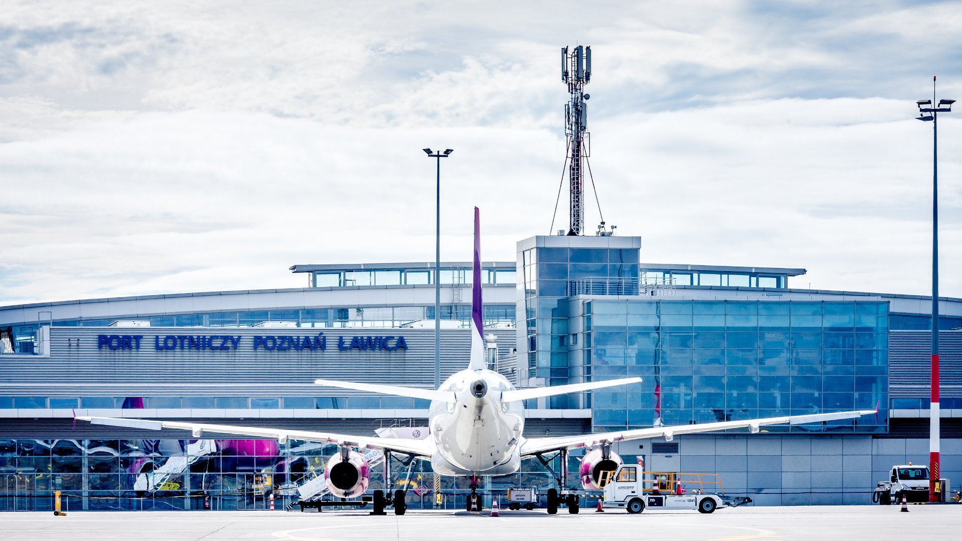 Port lotniczy Poznań-Ławica. Na pierwszym planie samolot z białym kadłubem i fioletowymi akcentami na ogonie oraz silnikach. Samolot zaparkowany na płycie lotniska, skierowany przodem w stronę kamery. Wokół samolotu znajdują się pojazdy obsługi lotniska oraz sprzęt. W tle budynek terminala z napisem "PORT LOTNICZY POZNAŃ ŁAWICA", wysoka wieża komunikacyjna i latarnie.