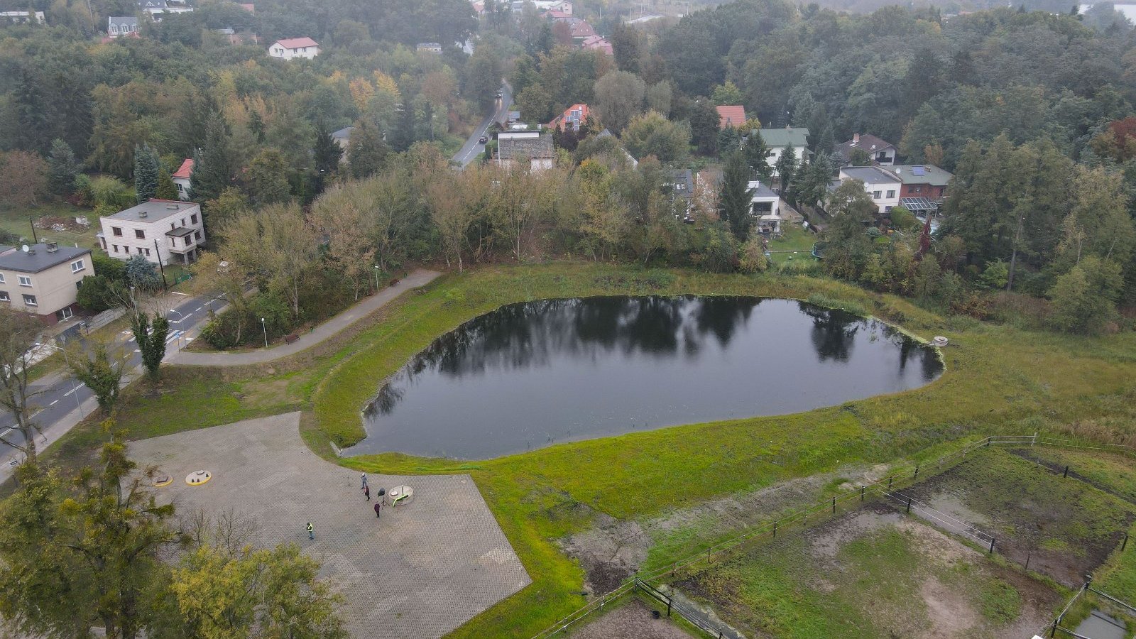 Zdjęcie przedstawia zbiornik retencyjny z lotu ptaka. Widać nam nim także budynki i drzewa.