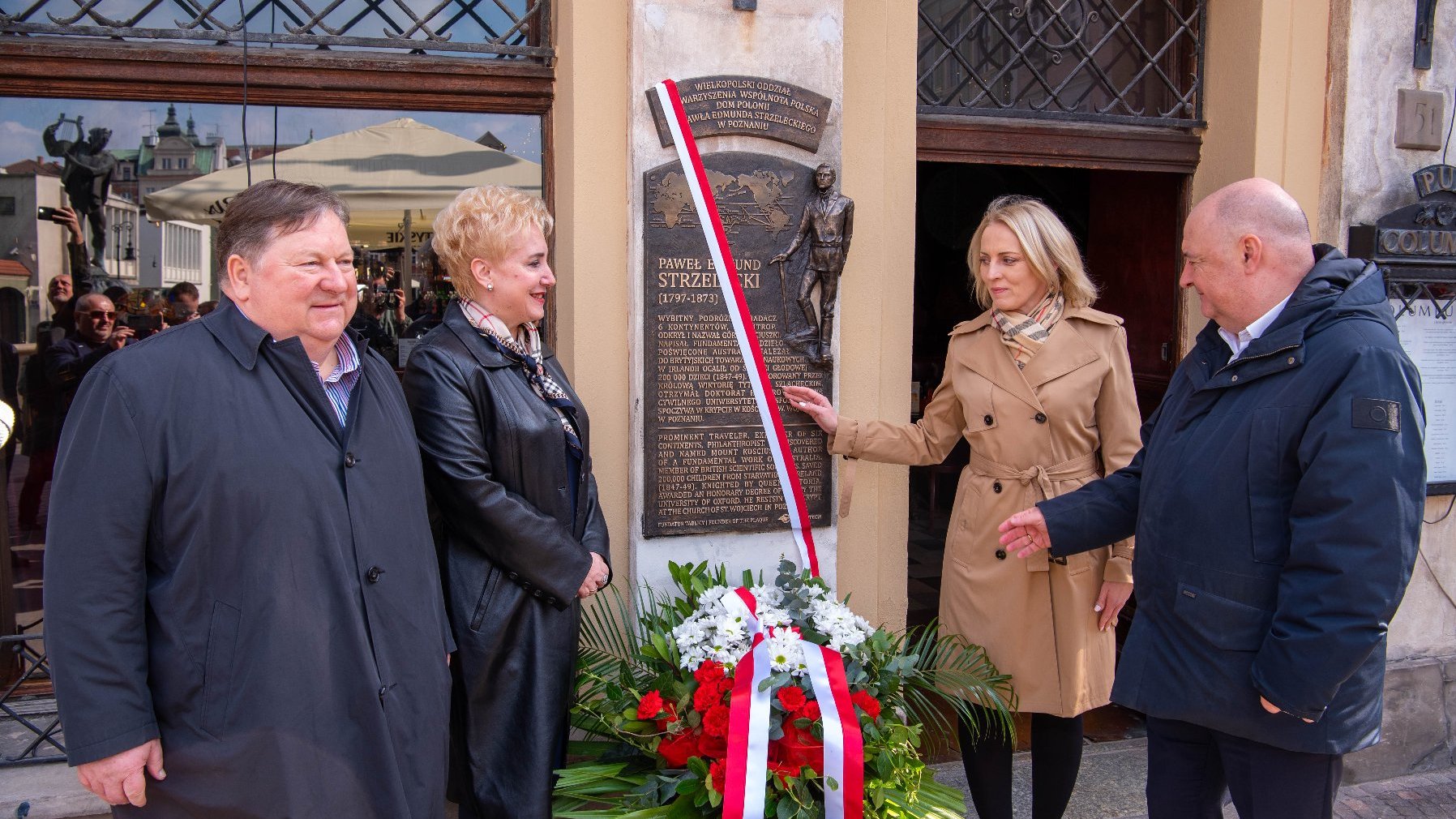 Czterech ludzi stoi przy tablicy pamiątkowej dedykowanej Pawłowi Edmundowi Strzeleckiemu, polskiemu odkrywcy i geologowi. Tablica zawiera jego nazwisko, datę życia (1797-1873) oraz informacje o jego osiągnięciach, w tym odkryciu Góry Kościuszki w Australii. Pod tablicą znajduje się duży biało-czerwony wieniec kwiatowy ozdobiony wstążką w kolorach polskiej flagi.