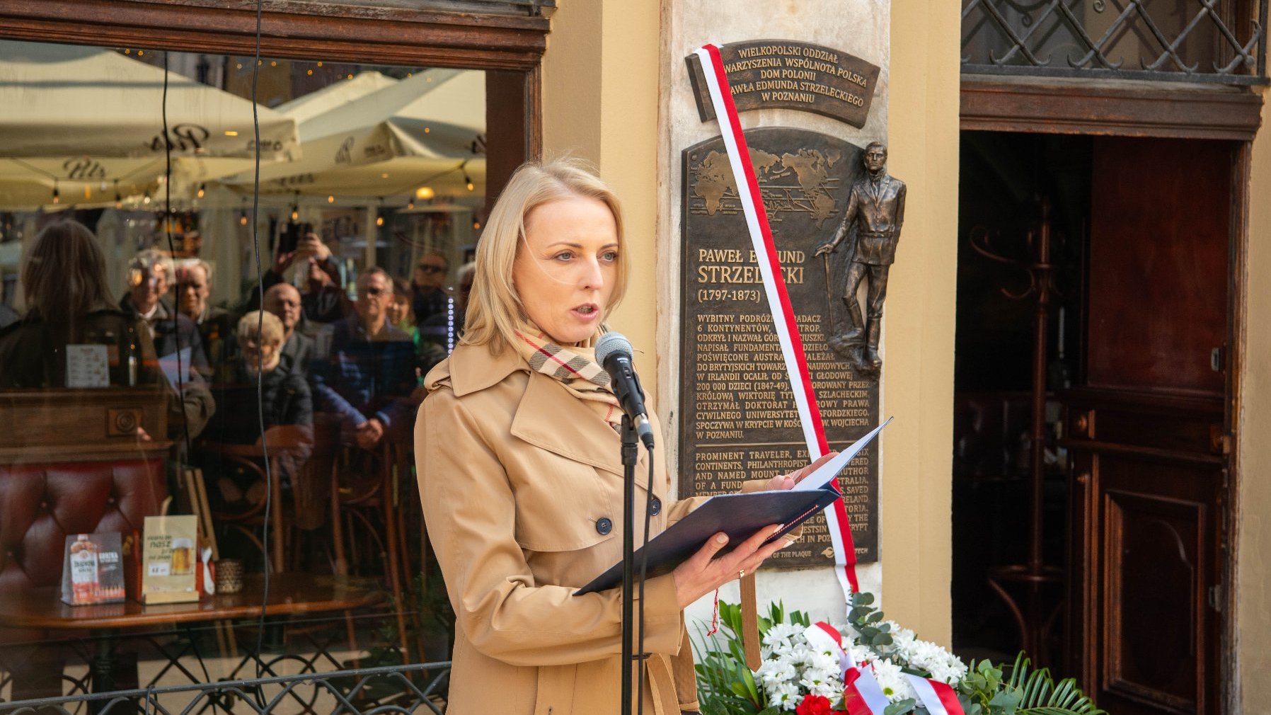 Kobieta przemawiająca do mikrofonu stoi przed tablicą pamiątkową poświęconą Pawłowi Edmundowi Strzeleckiemu (1797-1873), polskiemu podróżnikowi, geologowi i filantropowi. Tablica zawieszona na ścianie ozdobiona jest biało-czerwoną wstęgą.