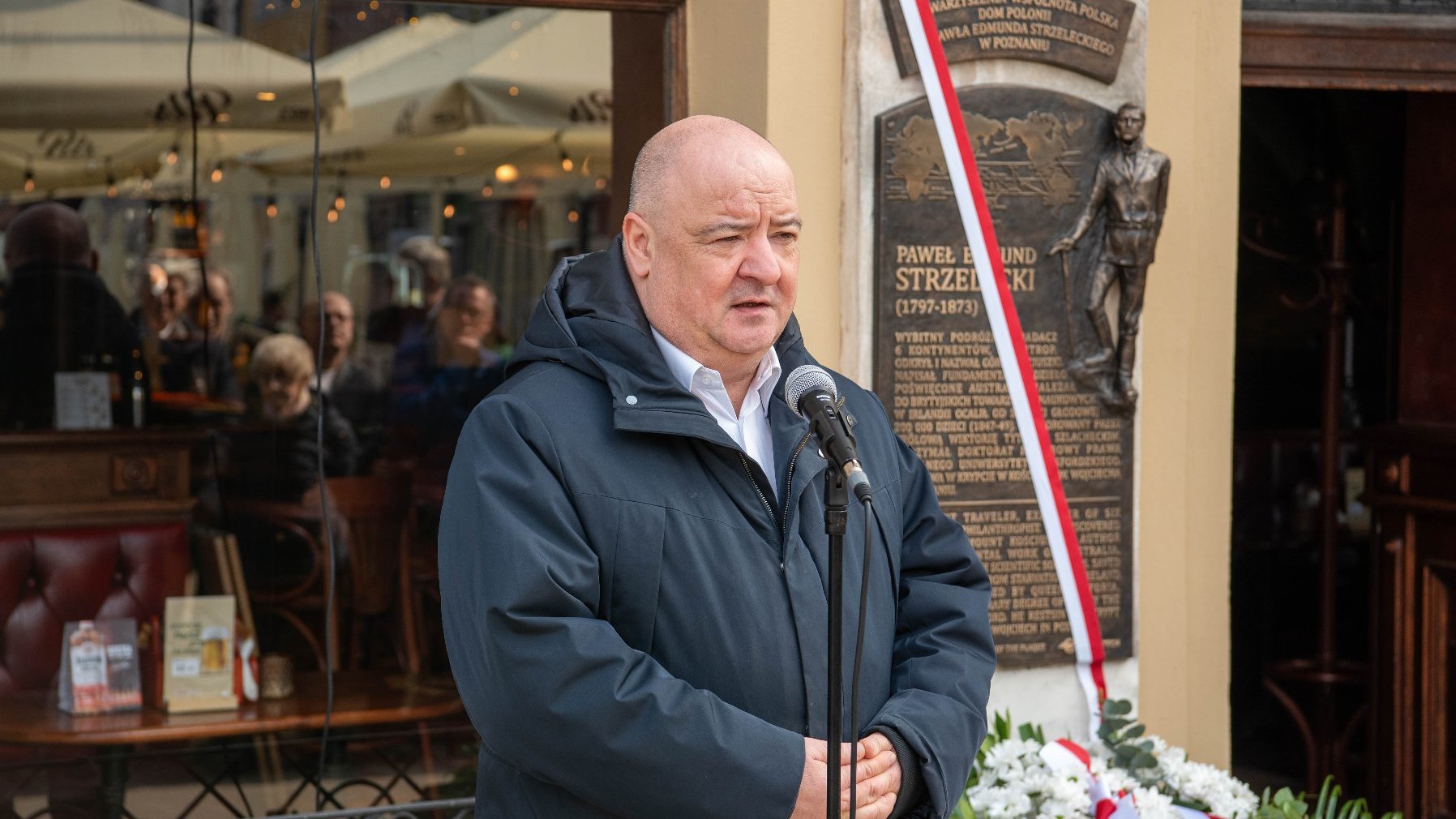 Osoba przemawia do mikrofonu, trzymając kartkę papieru, stoi przed budynkiem przy tablicy pamiątkowej. Tablica zawieszona na ścianie przedstawia postać mężczyzny oraz tekst opisujący jego dokonania. Ozdobiona jest wstęgą w kolorach polskiej flagi (biało-czerwona). U dołu tablicy znajduje się wieniec kwiatowy w kolorach białym i czerwonym, zdobiony wstążką w barwach polskiej flagi.